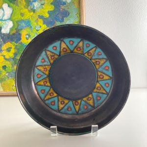 May include: A black ceramic plate with a blue, yellow, and orange geometric design. The design features a circle in the center with a sunburst pattern around it.