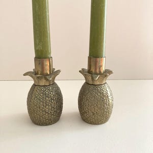 May include: A pair of brass candlestick holders shaped like pineapples, each featuring a textured, cross-hatched surface and a smooth metal socket at the top. Each holder contains a tall, slender, olive green taper candle.