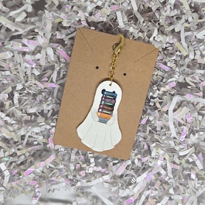 May include: A white ghost-shaped charm featuring a stack of colorful books held in its arms. The charm is attached to a gold-toned metal lobster clasp and chain, mounted on a rectangular brown card for display and secure storage.