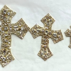 May include: Three gold-toned cross-shaped embellishments with clear rhinestones. The largest cross is about 5 inches tall, the middle cross is about 3 inches tall, and the smallest cross is about 2 inches tall.