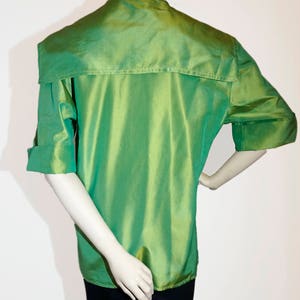 May include: A green, silky, short-sleeved shirt with a high collar and a large, folded collar. The shirt has a slightly gathered look and a sheen.