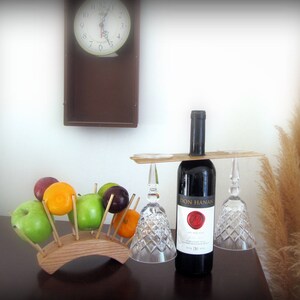Peut inclure: Nature morte avec une bouteille de vin rouge, deux verres à vin en cristal et un porte-fruits en bois rempli de pommes, d'oranges et de prunes. Une horloge est accrochée au mur en arrière-plan.
