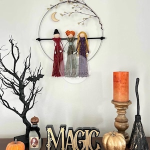 May include: A Halloween-themed wall hanging with three yarn dolls, a crescent moon, and a branch with white berries. The dolls are wearing red, green, and purple yarn dresses. The word "MAGIC" is displayed in gold letters on a black background.