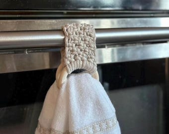 May include: A white kitchen towel with a beige trim hangs on a beige crocheted towel holder with wooden rings. The holder is attached to a silver oven handle.