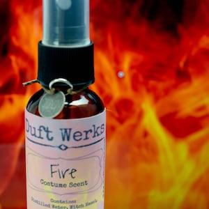 May include: A brown glass bottle with a black spray top and a pink label that reads "Duft Werks Fire Costume Scent". The label also says "Contains Distilled Water, Witch Hazel". The bottle is in front of a blurry background of orange and yellow flames.