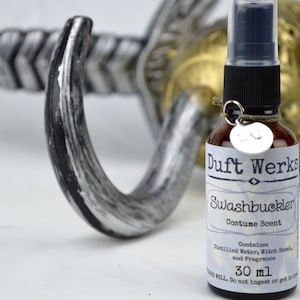 Puede incluir: Una pequeña botella de vidrio marrón con un pulverizador negro y una etiqueta blanca que dice "Duft Werks Swashbuckler Costume Scent". La etiqueta también incluye el texto "Contiene Agua Destilada, Hamamelis y Fragancia 30 ml".