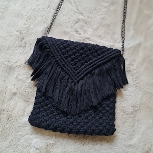 Puede incluir: Un bolso bandolera de crochet negro con ribete de flecos y una correa de cadena plateada.