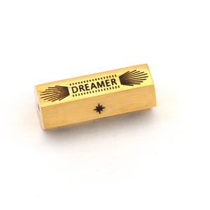 Op de afbeelding: Een goudkleurig, rechthoekig object met het woord "DREAMER" erop gedrukt. Het object heeft een ster- en zonnestraalontwerp. Het item staat tegen een witte achtergrond.