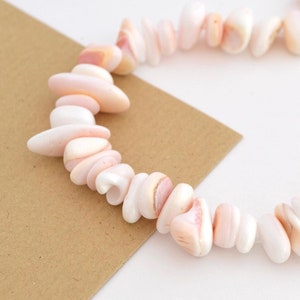 Op de afbeelding: Een streng roze en witte schelp kralen, geregen tot een ketting. De kralen zijn onregelmatig van vorm en hebben een natuurlijke, organische uitstraling.