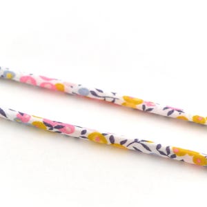 Op de afbeelding: Close-up van twee stoffen koorden met bloemenpatroon. De koorden zijn wit met een herhalend patroon van kleine roze, gele en blauwe bloemen met donkerblauwe bladeren. De koorden zijn enigszins onscherp.