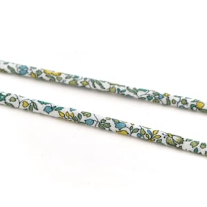 Può includere: Primo piano di due cordini in tessuto con motivo floreale. I cordini sono bianchi con un motivo ripetuto di piccole foglie e fiori blu, gialli e verdi. I cordini sono attorcigliati e sembrano essere di cotone.