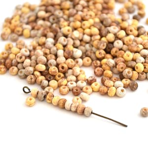 May include: A pile of small, round beads in beige, brown, and yellow hues, presented on a white background. Some beads are strung on a wire, demonstrating their stringing hole. Suitable for jewellery making or crafts.