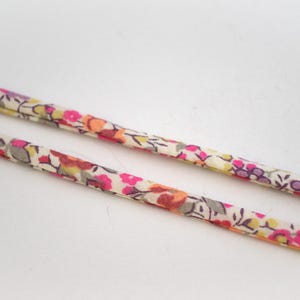 Puede incluir: Dos cordones finos recubiertos de tela con un patrón floral repetitivo en tonos rosa, naranja, amarillo y gris sobre un fondo color crema. Los cordones tienen forma cilíndrica y muestran una textura suave y consistente en toda su longitud.