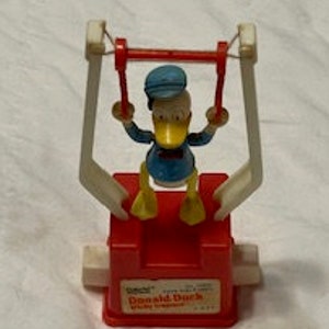 May include: A vintage Donald Duck toy in a red and white swing set. The toy features Donald Duck in a blue sailor suit and hat. The base of the toy is red with the words "Donald Duck" printed on it.