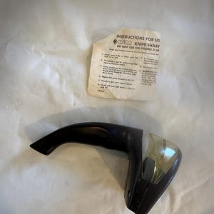 Puede incluir: Un afilador de cuchillos CUTCO negro con un mango curvo y un protector de cuchilla de pl&aacute;stico transparente. Se incluyen instrucciones de uso, con el texto "INSTRUCTIONS FOR US CUTCO KNIFE SHARF DO NOT USE ON DOUBLE D GE".