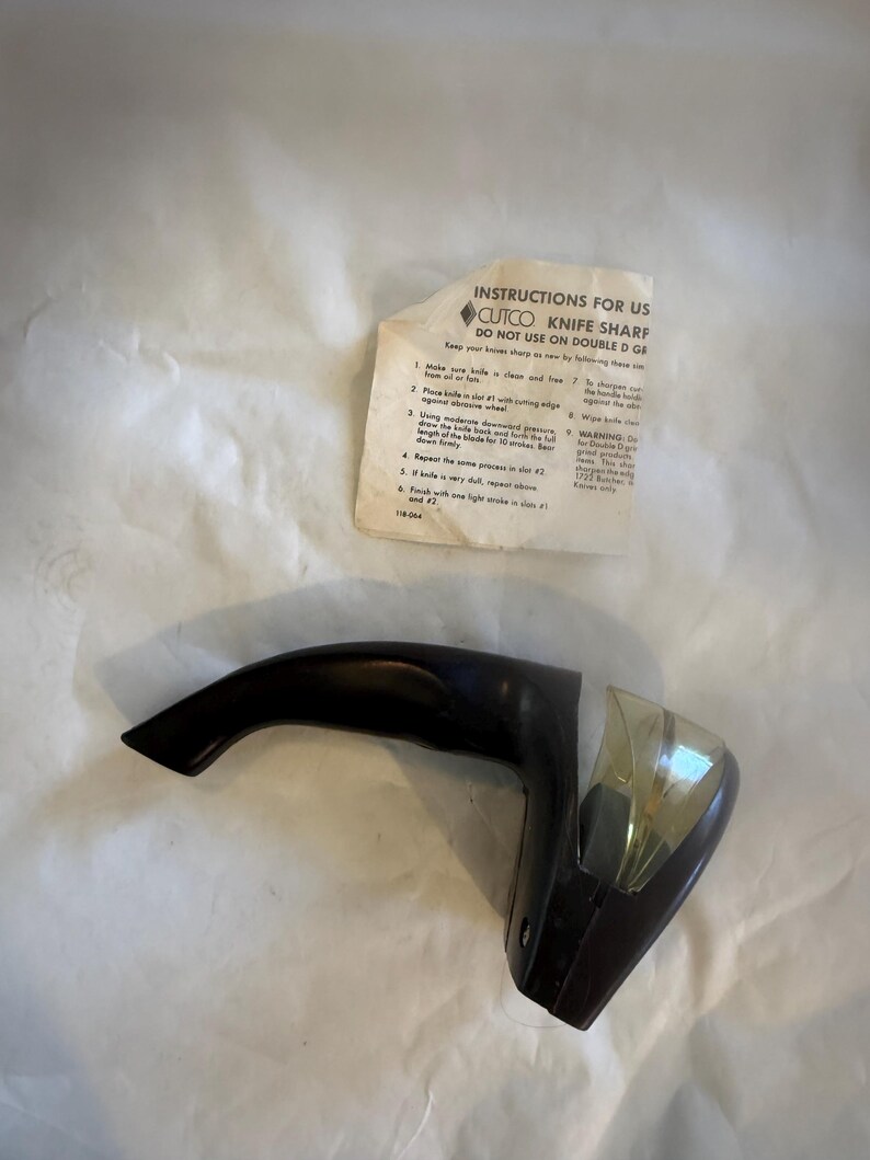 Puede incluir: Un afilador de cuchillos CUTCO negro con un mango curvo y un protector de cuchilla de pl&aacute;stico transparente. Se incluyen instrucciones de uso, con el texto "INSTRUCTIONS FOR US CUTCO KNIFE SHARF DO NOT USE ON DOUBLE D GE".