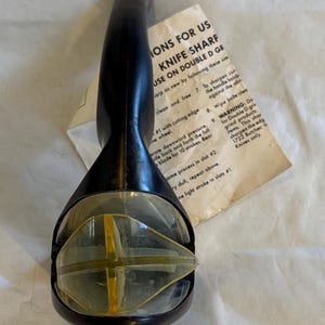 Puede incluir: Afilador de cuchillos negro y transparente con un elemento amarillo en forma de cruz. El afilador tiene un mango y una secci&oacute;n de pl&aacute;stico transparente. Las instrucciones est&aacute;n en papel, con el texto "KNIFE SHARP".