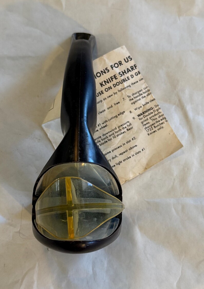 Puede incluir: Afilador de cuchillos negro y transparente con un elemento amarillo en forma de cruz. El afilador tiene un mango y una secci&oacute;n de pl&aacute;stico transparente. Las instrucciones est&aacute;n en papel, con el texto "KNIFE SHARP".