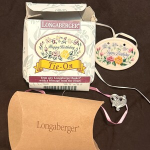 May include: A Longaberger Happy Birthday Tie-On set. The set includes a decorative box, a tag with floral accents, and a heart-shaped charm. The box and tag feature the words "Happy Birthday". The set is designed to trim a Longaberger basket.
