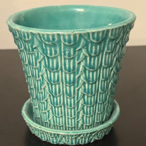 May include: A turquoise ceramic plant pot with a matching saucer, featuring a raised, repeating scale-like texture across the exterior surface. The pot has a wide, open circular rim and a tapered body that sits securely within the circular base tray.