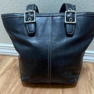 May include: A black leather tote bag featuring two shoulder straps with silver-toned metal buckles. The bag has a vertical seam down the center and white contrast stitching along the edges, the straps, and the buckle attachments.