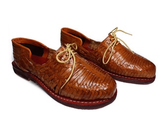 Könnte beinhalten: Ein Paar braune Ledersandalen mit geflochtenen Lederriemen und Schnürsenkeln. Die Sandalen haben ein rotes Nähtdetail um die Sohle.