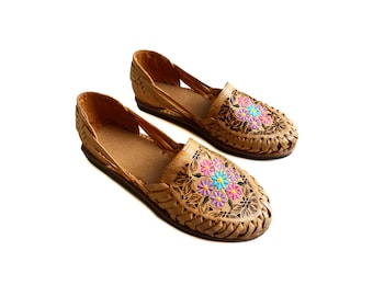 Könnte beinhalten: Ein Paar sandfarbene Ledersandalen mit Blumenstickerei in Rosa, Blau und Gelb. Die Sandalen haben ein geflochtenes Riemen-Design und eine spitze Zehenpartie.
