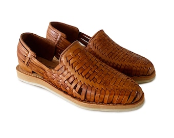 May include: A pair of tan huarache shoes featuring a woven leather upper with a closed toe and an open heel design. The shoes have a flat, light-colored sole and a complex, interlaced pattern across the top and sides of the footwear.