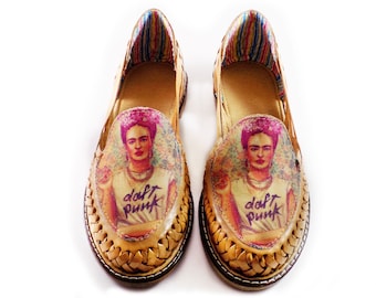 Pode incluir: Um par de sandálias de couro castanho com um design tecido. As sandálias apresentam uma imagem impressa de Frida Kahlo usando uma camisa branca com o texto "Daft Punk" em roxo. As sandálias têm um forro listrado colorido.