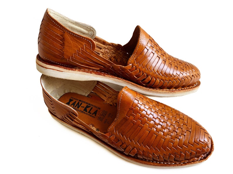 K&ouml;nnte beinhalten: Ein Paar braune Ledersandalen mit gewebtem Design. Die Sandalen haben eine wei&szlig;e Sohle und werden von der Marke "Xan-Kla" hergestellt.