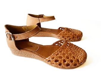 May include: Tan leather sandals with a woven design and an ankle strap. The sandals have a wedge heel.