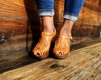 Puede incluir: Un par de sandalias de plataforma de cuero marrón con correas tejidas y un tacón grueso. Las sandalias las lleva una persona de pie sobre una superficie de madera.