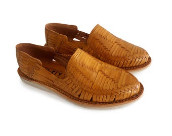 May include: A pair of tan leather woven huarache sandals. The shoes have a slip-on design with an open top and a woven pattern across the top and sides. The soles are a light color.