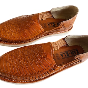 K&ouml;nnte beinhalten: Ein Paar braune Ledersandalen mit gewebtem Muster. Die Sandalen haben einen Lederriemen &uuml;ber dem Fu&szlig;r&uuml;cken und eine Ledersohle. Der Markenname "YAN-KLA" ist auf der Innenseite der Sandalen sichtbar.