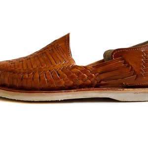 K&ouml;nnte beinhalten: Ein Paar braune Ledersandalen mit gewebtem Design. Die Sandalen haben eine wei&szlig;e Sohle und eine geschlossene Zehenpartie.