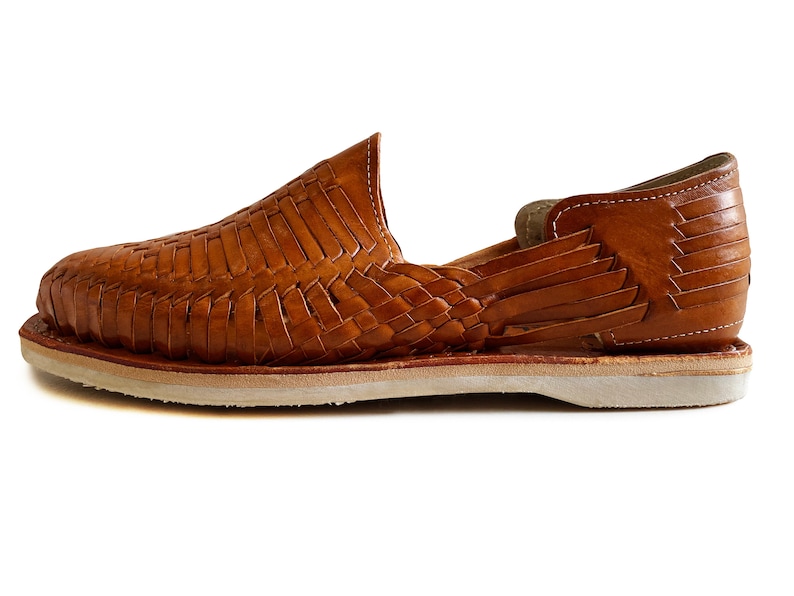 K&ouml;nnte beinhalten: Ein Paar braune Ledersandalen mit gewebtem Design. Die Sandalen haben eine wei&szlig;e Sohle und eine geschlossene Zehenpartie.