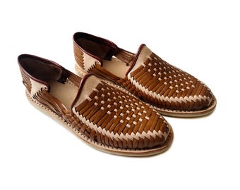 Könnte beinhalten: Ein Paar Huarache-Schuhe mit einem geflochtenen Muster aus braunen und beigen Lederstreifen. Die Schuhe haben eine geschlossene Zehenpartie, ein offenes Seitendesign und eine flache Sohle mit einem dunkelbraunen Lederbesatz am Schaft.