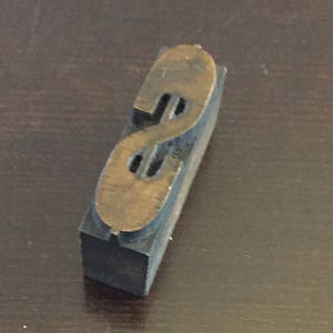 以下が含まれることがあります： ドル記号の形をした金属製の文字 'S' が付いた、ビンテージの木製印刷ブロックです。