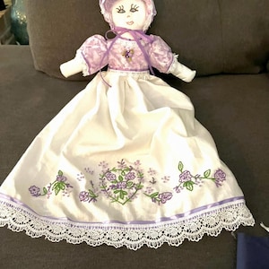 Könnte beinhalten: Handgefertigte Puppe mit Häubchen und weißem Kleid. Das Kleid ist mit lila Blumenstickerei und einer Spitzenborte verziert. Mieder und Ärmel der Puppe sind aus passendem lila Stoff gefertigt.