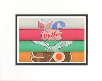 May include: A stack of five folded t-shirts with different designs. The top shirt is white with red, white, and blue stars and the number 6. The second shirt is pink with a baseball and the text "Phillies". The third shirt is green with a white eagle. The fourth shirt is orange with a brown circle and a smaller orange circle inside. The bottom shirt is brown with a white circle and a smaller orange circle inside.