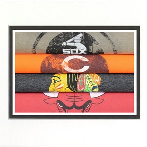 May include: A stack of four t-shirts with different sports team logos. The top shirt is gray with the word "SOX" on it. The second shirt is orange with a white "C" on it. The third shirt is gray with a Native American headdress on it. The bottom shirt is red with a black bull's head on it.