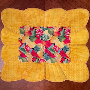 May include: A yellow quilted table runner with a scalloped edge features a central panel with a colorful fruit design. The fruit design includes watermelon, lemons, limes, and oranges.