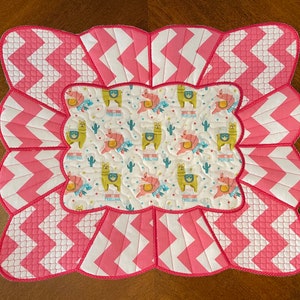May include: A quilted placemat with a scalloped edge. The center features a pattern of llamas, cacti, and the text "PRISCILLAMA" on a white background. The outer border has a pink and white chevron pattern.
