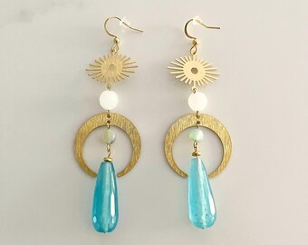 Könnte beinhalten: Ein Paar goldfarbene Ohrringe mit einem himmlischen Design. Jeder Ohrring hat eine sonnenförmige Oberseite, eine weiße Perle, einen goldenen Halbmond, eine hellgrüne Perle und einen tropfenförmigen blauen Edelstein. Die Ohrringe werden auf einer weißen Oberfläche präsentiert.