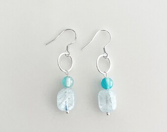 Puede incluir: Pendientes de plata con un colgante de piedra preciosa azul claro y azul oscuro. Los pendientes tienen un pequeño aro de plata en la parte superior.