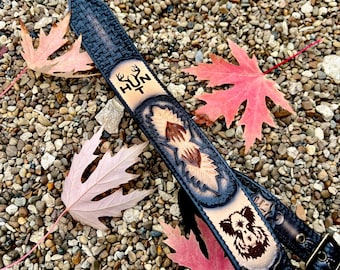 May include: A black leather rifle sling with embossed designs and the word "HUNT" with deer antlers. Features a bear illustration and initials "GT". The sling is decorated with a floral pattern and a buckle.