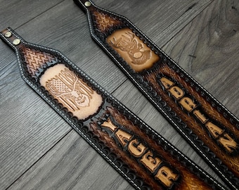 May include: Two brown leather gun slings with black stitching. One sling has the name "REGAN YA" embossed on it and the other has the name "ADRIAN" embossed on it. Both slings feature a deer head design.