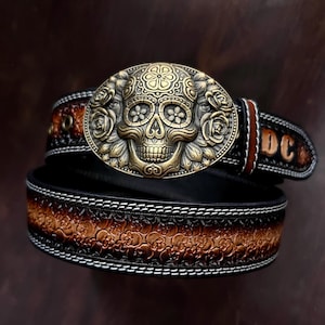 May include: A black leather belt with a decorative brass skull buckle. The buckle features a detailed skull design with floral accents. The belt has a brown embossed pattern and the initials "DC" are visible.