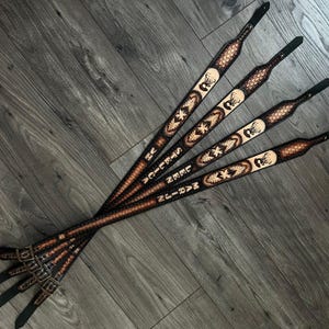 May include: Four brown leather guitar straps with a deer head design and personalized text. The straps are laid out in a row on a light brown wood surface.