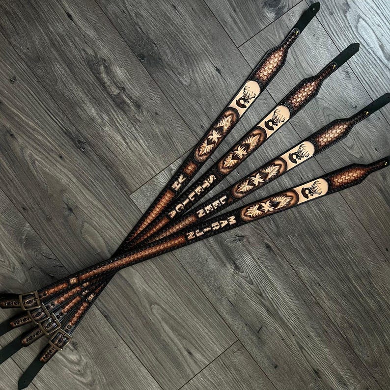 May include: Four brown leather guitar straps with a deer head design and personalized text. The straps are laid out in a row on a light brown wood surface.